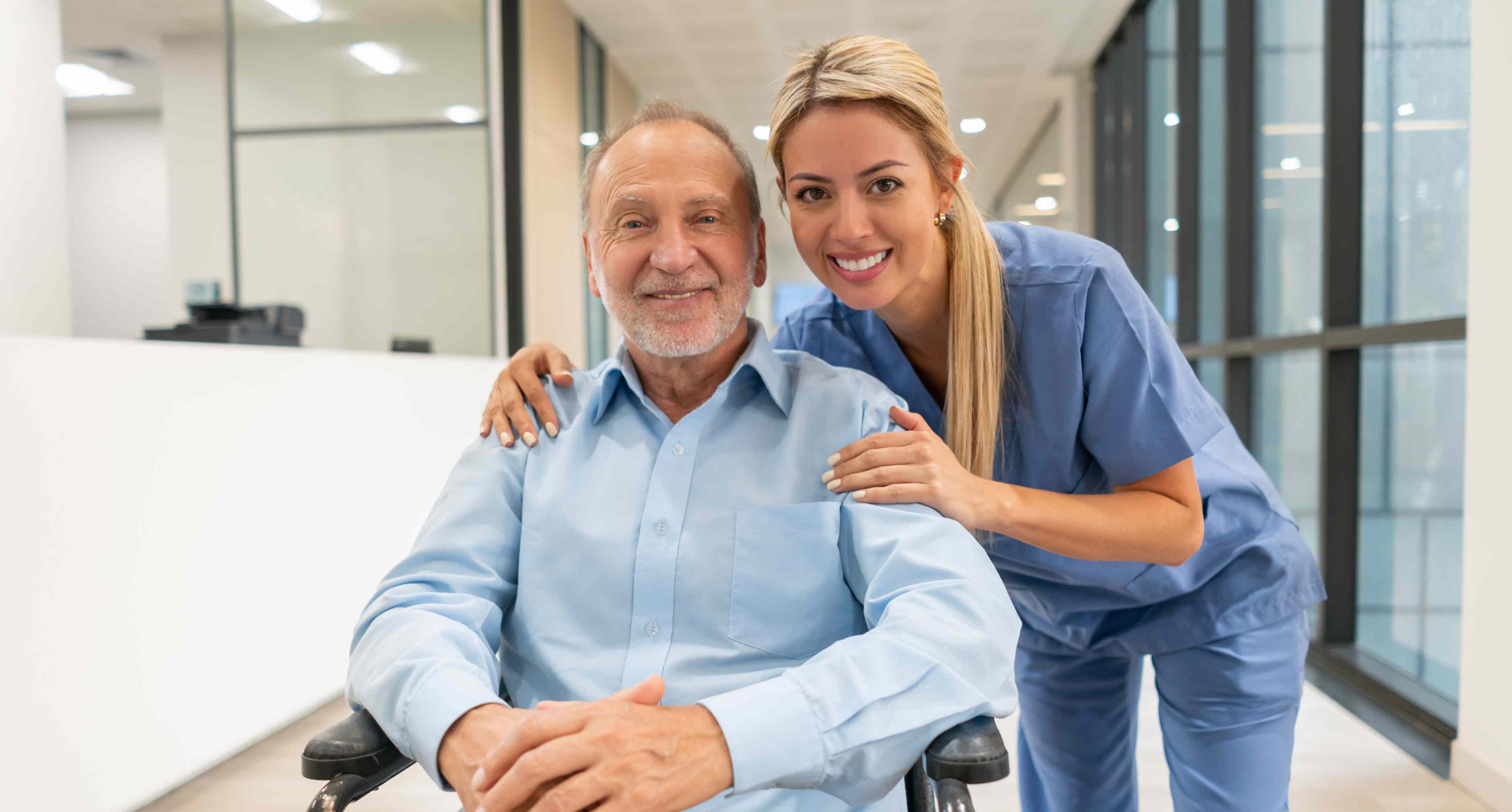 Smiling elderly man seated in a wheelchair with a healthcare professional standing beside him, both looking confident and supported in a clinical setting.