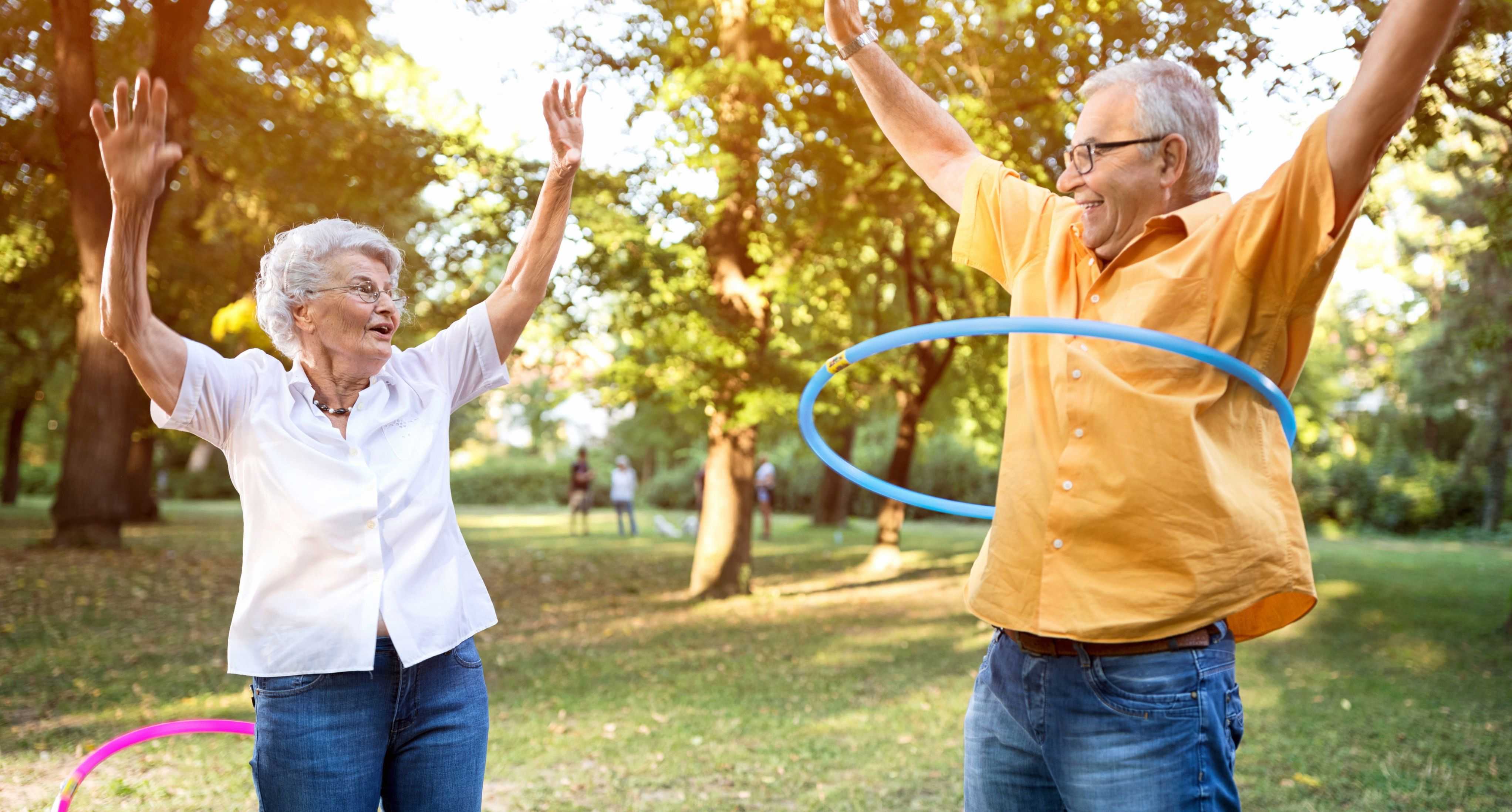 Smiling senior couple exercising outdoors in a park, raising their arms while using hula hoops, enjoying active and playful movement.