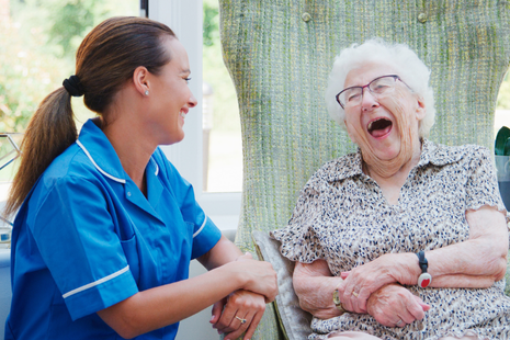 This image shows a smiling older man sitting beside a healthcare professional in scrubs, who has her arm gently around his shoulder. Both are looking at the camera warmly, conveying a sense of trust, care, and companionship.