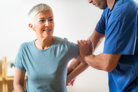 an older woman smiling while a healthcare professional assists her with a physical therapy or rehabilitation exercise.
