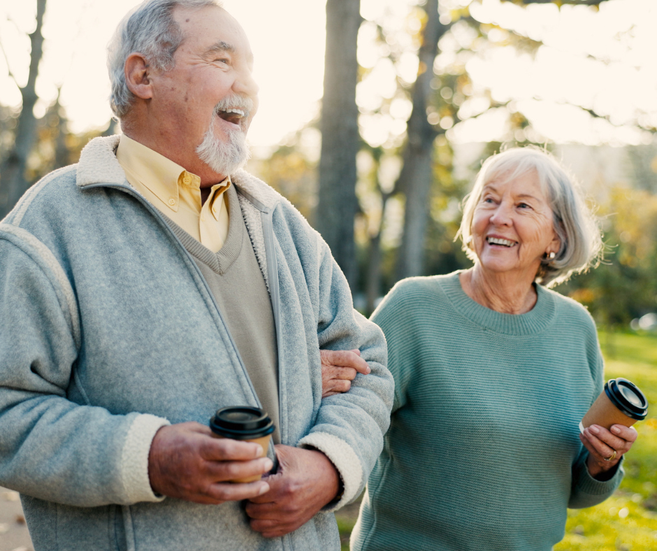A cheerful caregiver in scrubs sits beside an older man on a couch, both engaged and smiling while interacting with a tablet. The scene highlights companionship, digital engagement, and supportive aged care in a comfortable, home-like setting.